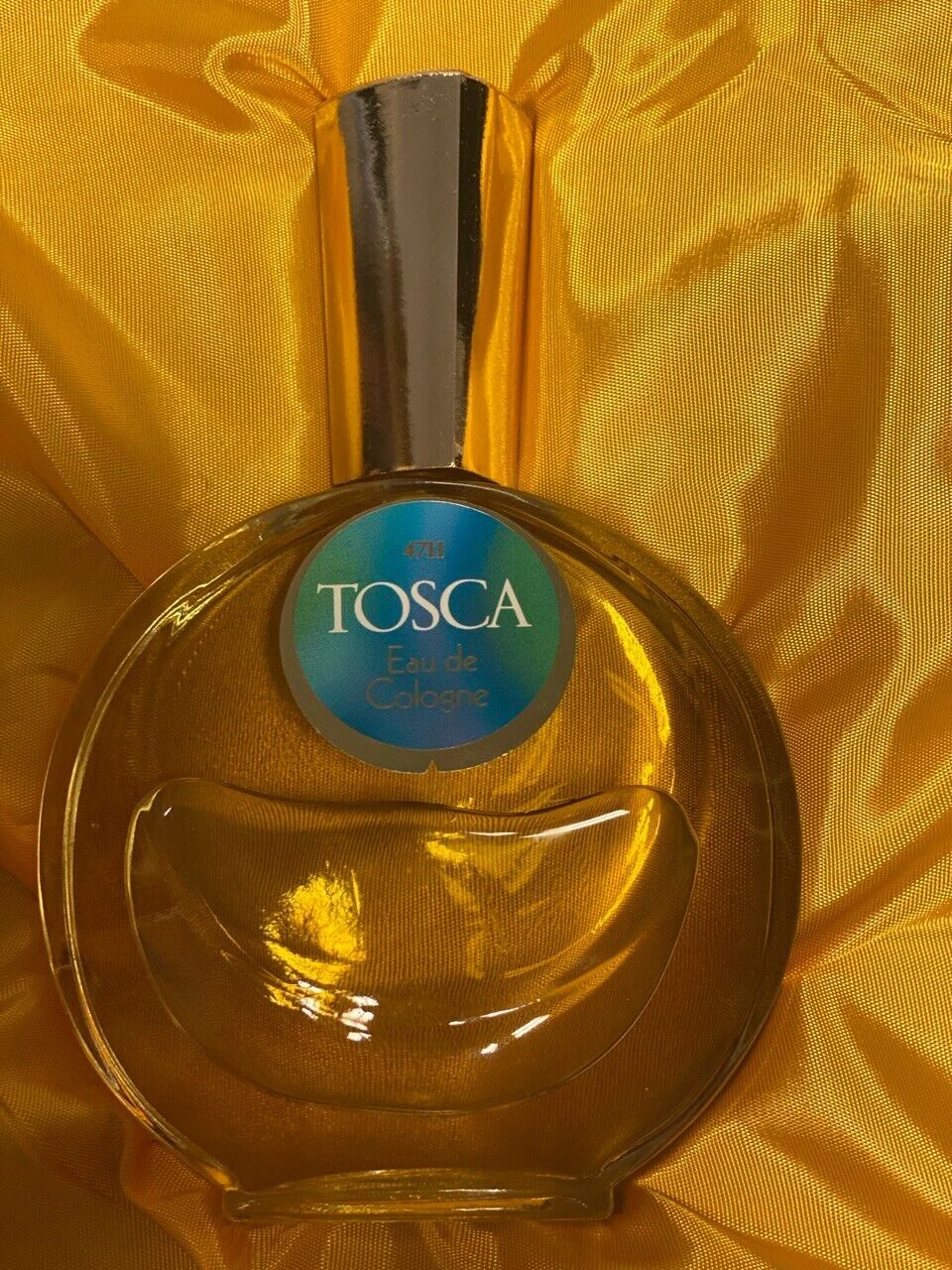 Ein rundliches, bernsteinfarbenes Parfumflakon von „4711 Tosca Eau de Cologne“ steht auf glänzendem, goldgelbem Stoff. Der silberne Verschluss reflektiert das Licht, während das türkis-blaue Etikett mit goldener Schrift zentral auf der Flasche platziert ist.