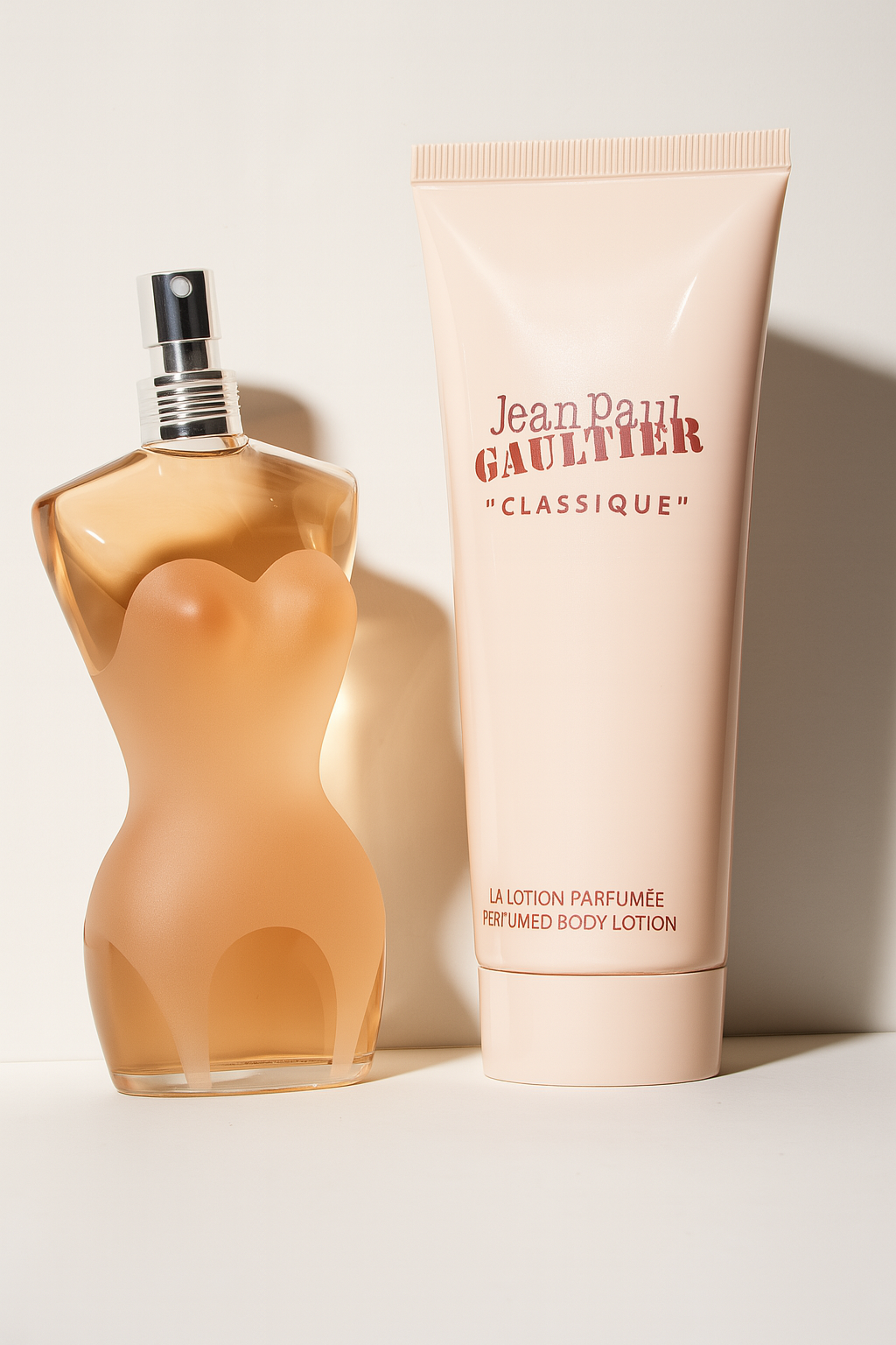 Ein leicht seitlich gekipptes Bild zeigt das Jean Paul Gaultier Classique Duft-Set. Links steht ein elegant geformtes, torsoförmiges Eau de Toilette Flakon aus mattem Glas mit glänzendem Sprühkopf. Rechts daneben eine aufrecht stehende, glänzende Tube der Bodylotion mit dem typischen Markenaufdruck. Beide Produkte werfen einen sanften, natürlichen Schatten auf einen neutralen Hintergrund.