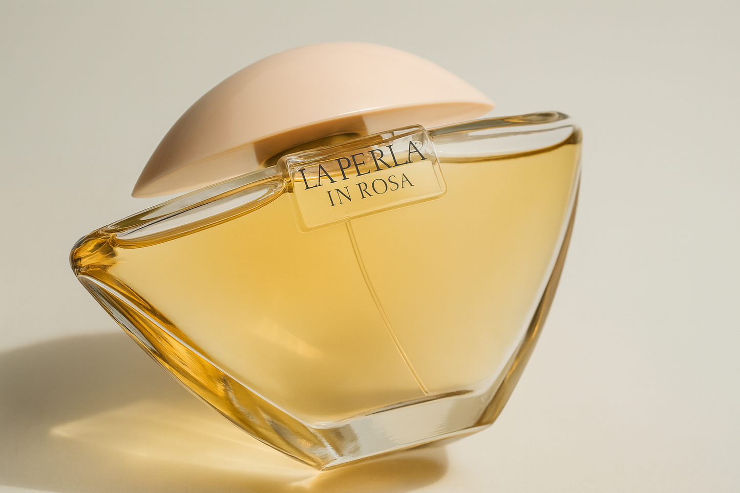 Ein leicht nach hinten gekipptes, elegantes La Perla „In Rosa“-Parfumflakon. Der Glasflakon enthält eine goldene Flüssigkeit, wird von einem zarten, roséfarbenen Deckel verschlossen und wirft einen weichen Schatten auf einen neutralen Hintergrund.