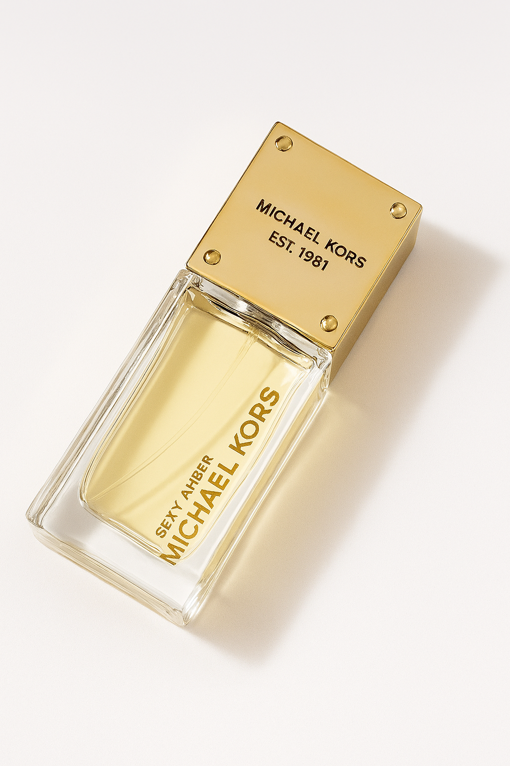 Ein geneigter Flakon des Parfums „Sexy Amber“ von Michael Kors liegt auf einem hellen, neutralen Untergrund. Der goldene Deckel und das klare Glas mit bernsteinfarbener Flüssigkeit werfen einen sanften Schatten und verleihen dem Bild Tiefe und Eleganz.