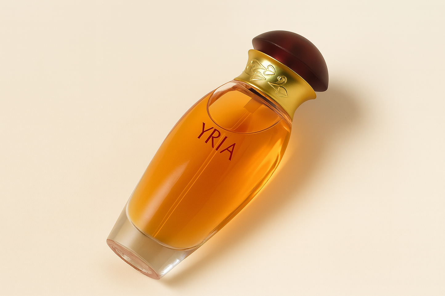 Ein elegantes Parfumflakon mit bernsteinfarbener Flüssigkeit liegt leicht geneigt auf einem cremefarbenen Hintergrund. Das klare Glas zeigt die rote Aufschrift „YRIA“. Der Flakon ist mit einem goldfarbenen Hals verziert, der florale Gravuren trägt. Die runde Kappe ist bordeauxfarben mit glänzender Oberfläche. Ein weicher Schatten sorgt für Tiefe.