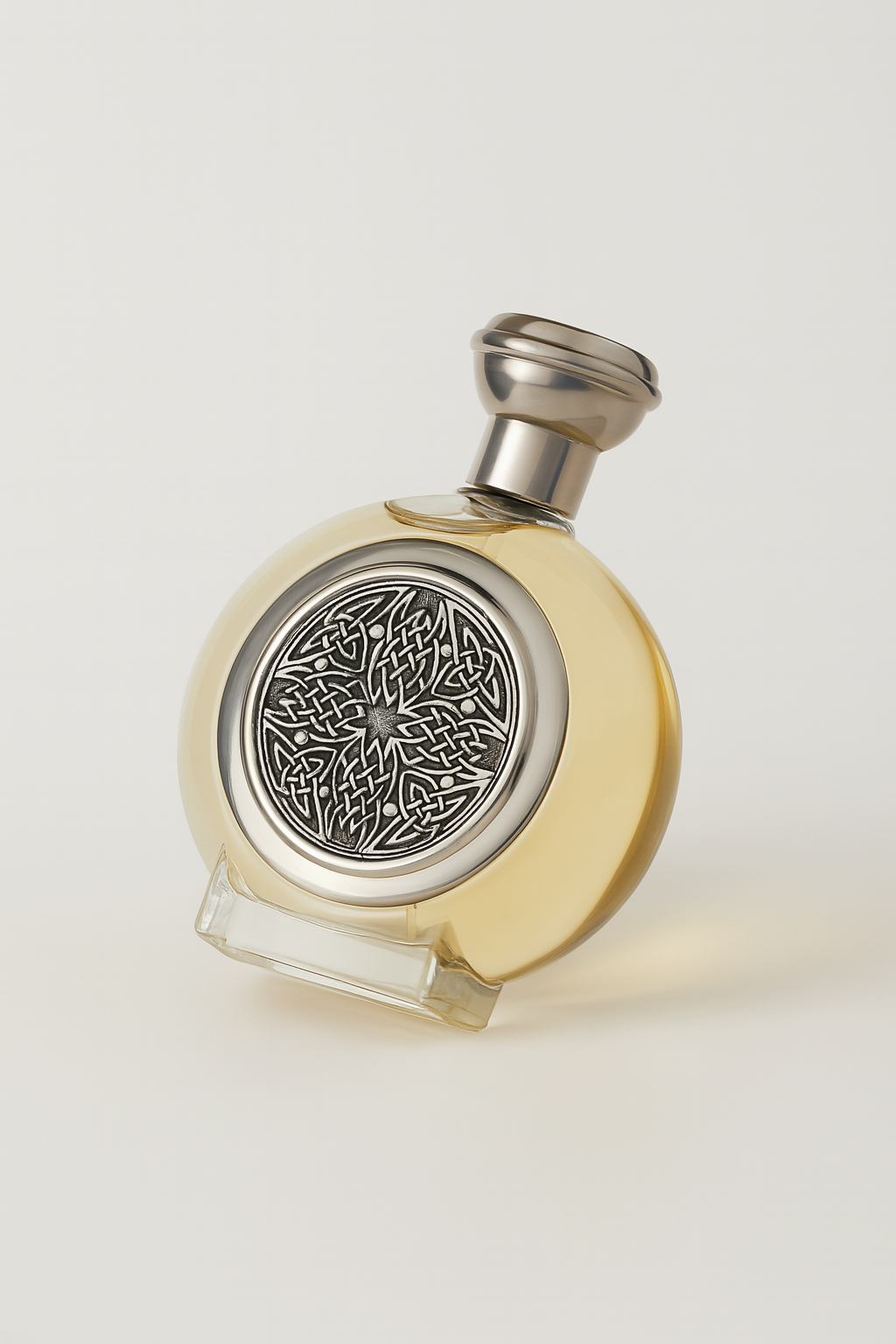 Ein rundes Parfumflakon von Boadicea the Victorious mit einem silbernen, verzierten keltischen Muster auf der Vorderseite, gefüllt mit hellgoldener Flüssigkeit. Es steht leicht nach hinten gekippt auf einem neutralen Hintergrund mit weichem Schattenwurf.