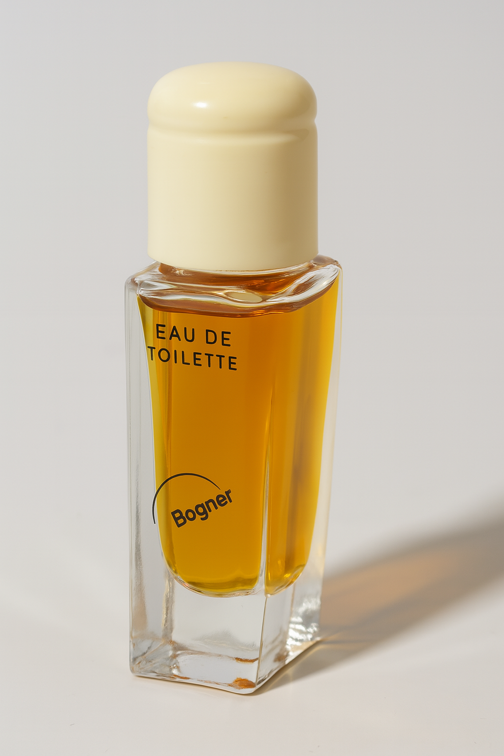 Ein rechteckiges Eau de Toilette-Flakon von Bogner mit transparenter Glasfront, gefüllt mit bernsteinfarbener Flüssigkeit, und einem cremefarbenen Verschluss. Das Flakon steht leicht zur Seite gekippt vor einem neutralen Hintergrund und wirft einen weichen Schatten.