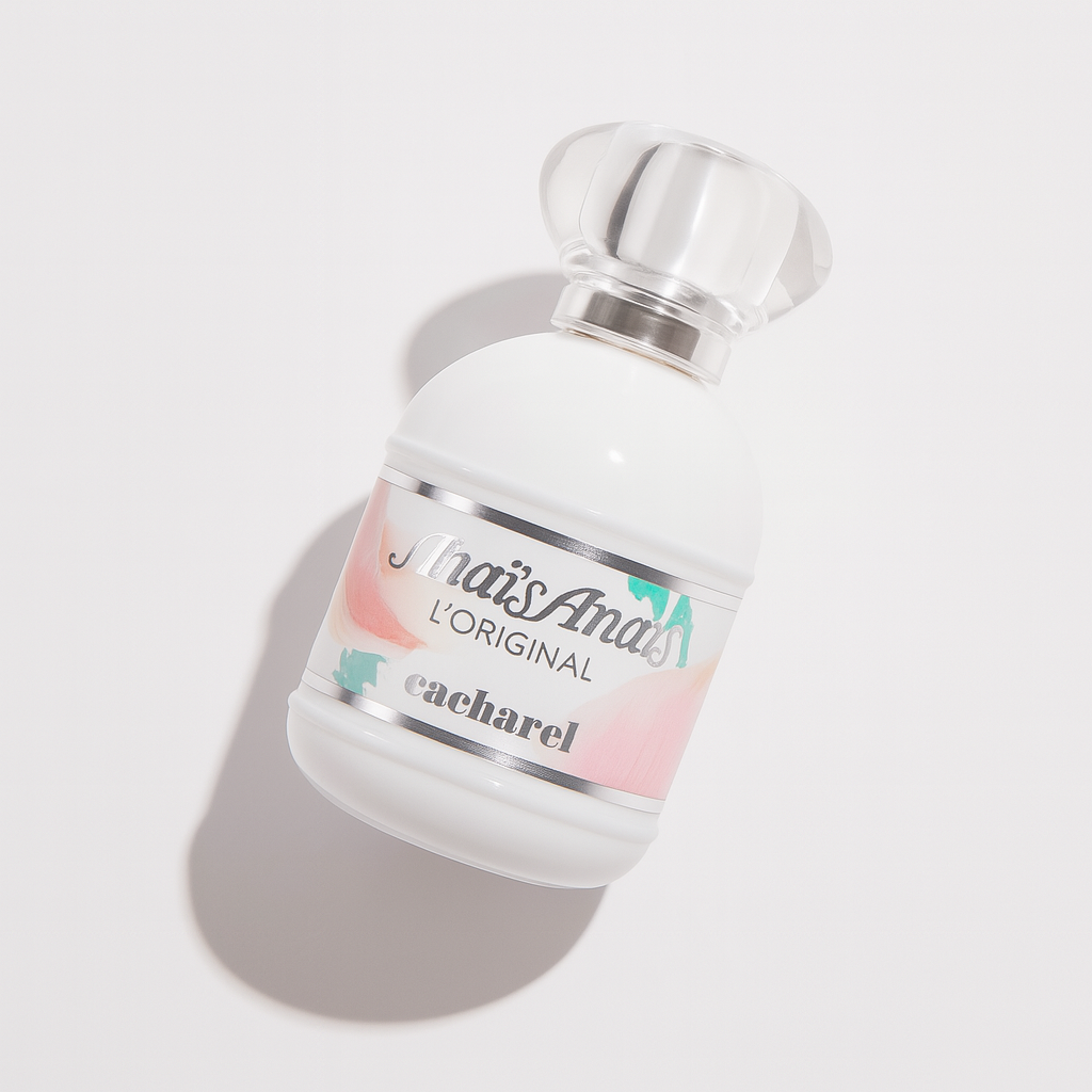 Ein weißes, rundliches Parfümflakon von Cacharel Anaïs Anaïs L'Original liegt leicht nach hinten gekippt auf einer hellen Fläche. Das Etikett zeigt sanfte Pastelltöne in Rosa und Pfirsich mit silbernen Akzenten, während der silberne Verschluss glänzt und ein weicher Schatten nach hinten fällt.