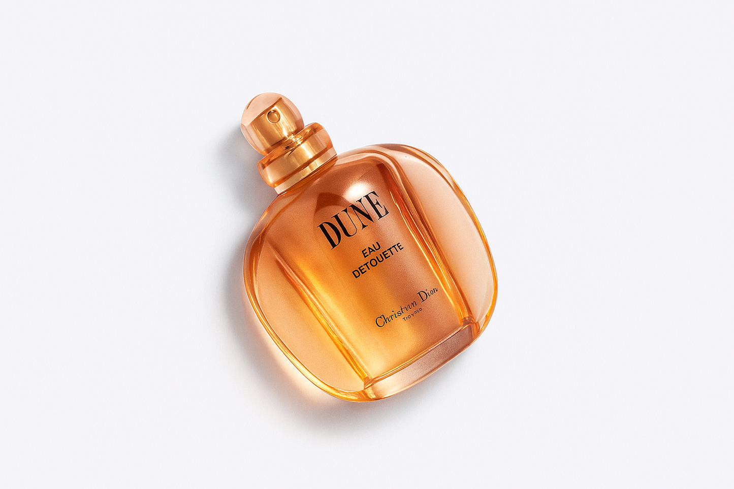 Ein schräg fotografiertes Parfumflakon von "DUNE Eau de Toilette" von Christian Dior, mit einer eleganten, abgerundeten Glasform in warmem Bernsteinfarbton. Die Aufschrift ist klar lesbar, der Hintergrund ist neutral weiß und das Flakon wirft einen sanften, natürlichen Schatten.
