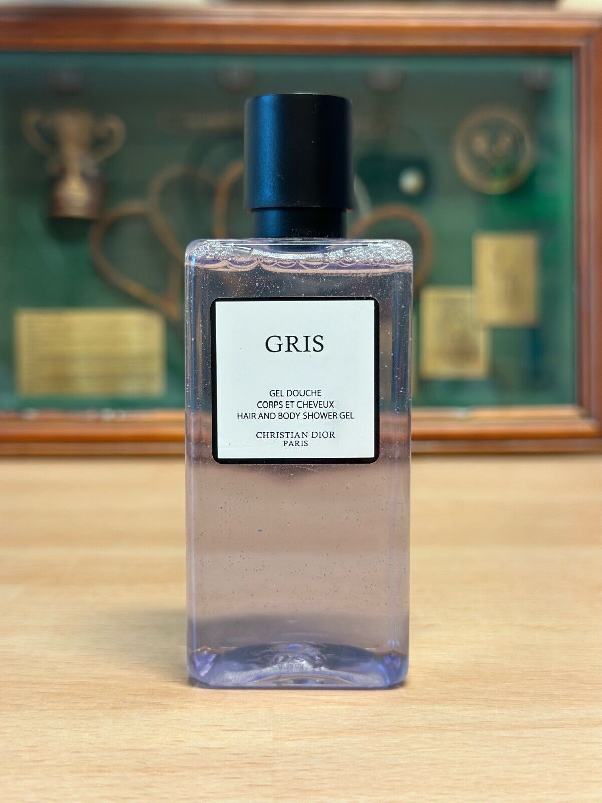 Dior Gris Duschgel Flasche steht auf einer hölzernen Fläche. Im Hintergrund ist leicht verschwommen ein Glaskaten mit Tennis-Utensilien zu erkennen.