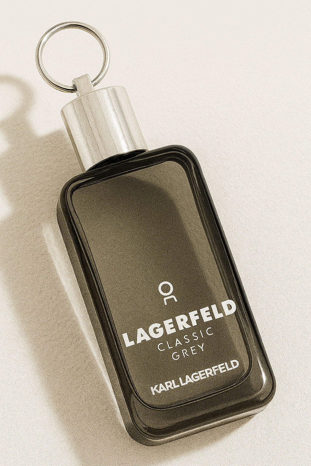 Ein rechteckiges, graues Parfumflakon mit silbernem Sprühkopf und rundem Metallring liegt leicht nach hinten gekippt auf einer neutralen, hellen Oberfläche. Die Aufschrift „Lagerfeld Classic Grey“ ist auf der Vorderseite klar lesbar. Das Flakon wirft einen sanften, natürlichen Schatten.