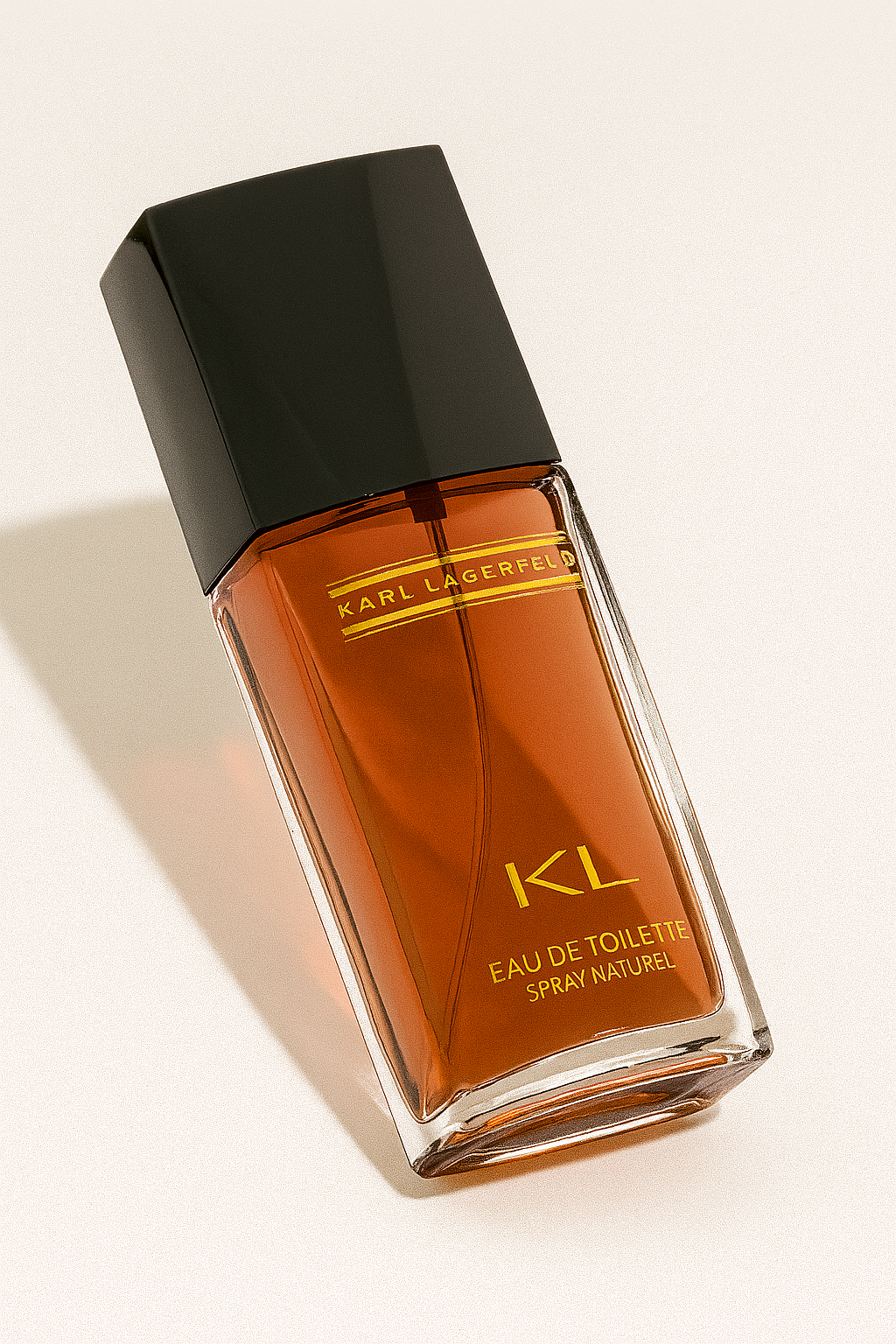 Eine seitlich gekippte Flasche des Parfums „KL Eau de Toilette Spray Naturel“ von Karl Lagerfeld, aus klarem Glas mit bernsteinfarbener Flüssigkeit und schwarzer Verschlusskappe, steht auf einem neutralen hellen Hintergrund mit weichem, natürlichem Schatten.