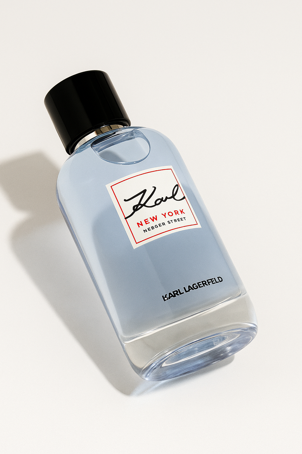 Ein eleganter, seitlich gekippter Flakon des Parfums „Karl Lagerfeld New York Mercer Street“ liegt auf einem neutralen, hellen Hintergrund. Das Glasfläschchen enthält eine hellblaue Flüssigkeit und hat einen schwarzen Deckel sowie eine weiße Etikette mit roter und schwarzer Aufschrift. Ein weicher Schatten und dezente Lichtreflexionen verleihen dem Bild Tiefe.