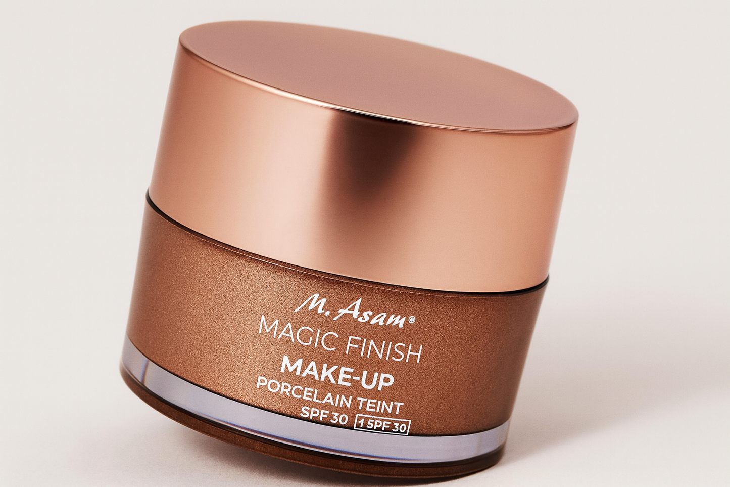 Eine Dose der M. Asam Magic Finish Make-Up Porcelain Teint SPF 30 liegt leicht nach hinten geneigt auf einem neutralen Hintergrund. Der bronzefarbene Tiegel mit roségoldenem Deckel reflektiert sanft das Licht, während die weiße Aufschrift klar sichtbar ist. Ein weicher Schatten betont die elegante Rundung und Tiefe.
