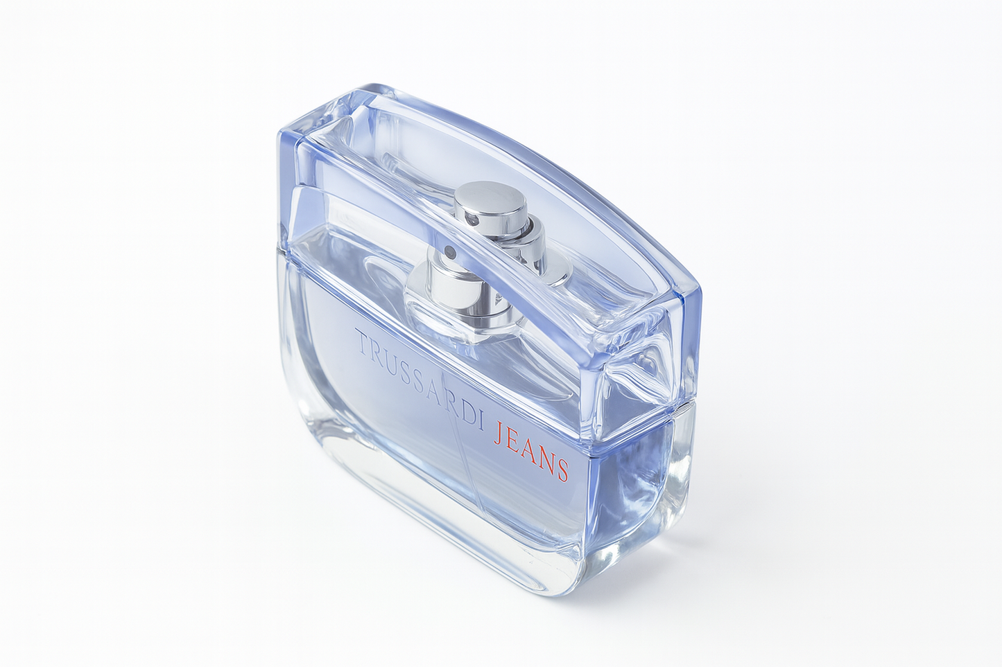 Ein schräg fotografierter Flakon des Parfums "Trussardi Jeans" mit transparent-blauem Glasdesign, silbernem Sprühkopf und roter Aufschrift, vor neutralweißem Hintergrund.