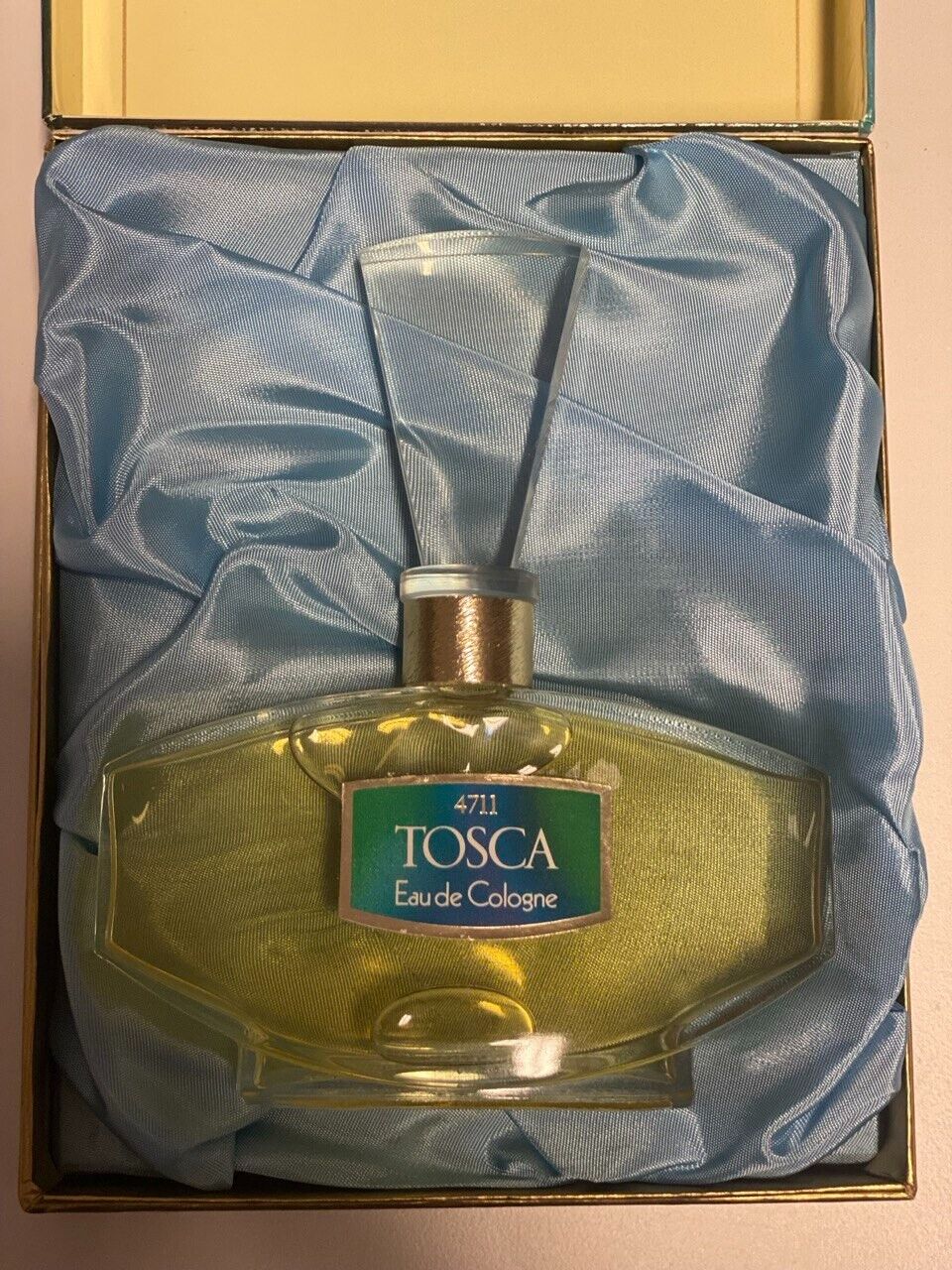 Ein elegantes, eckig geschwungenes Flakon von „4711 Tosca Eau de Cologne“ mit goldener Verschlusskappe liegt in einer Geschenkbox auf hellblauem Satin. Das türkis-blaue Etikett mit silberner Umrandung und weißer Schrift ist mittig auf der Flasche angebracht.