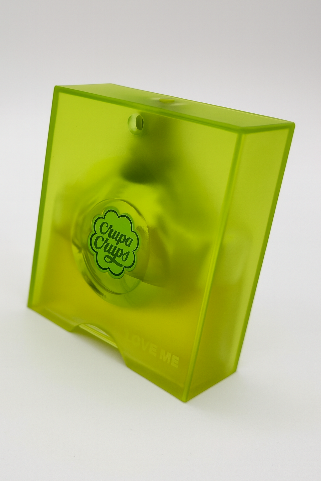 Ein nach hinten geneigtes, quadratisches Parfumflakon von Chupa Chups „Love Me“ in leuchtendem, transparentem Neongrün. Das Flakon zeigt mittig das Chupa-Chups-Logo in Dunkelgrün. Der Hintergrund ist neutral weiß, ein weicher Schatten fällt nach hinten.