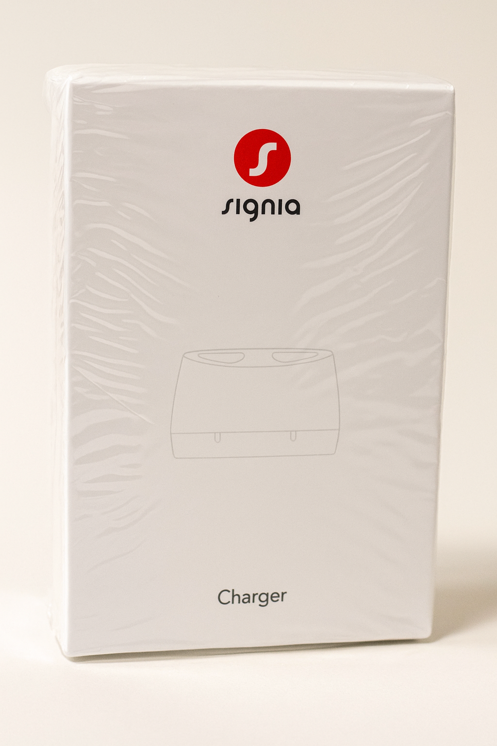 Eine rechteckige, in transparenter Folie eingeschweißte Produktverpackung des Signia Chargers für Hörgeräte. Die weiße Schachtel steht leicht nach hinten geneigt auf neutralem Hintergrund. Auf der Vorderseite ist das rote Signia-Logo, der Markenname „Signia“ sowie eine graue Skizze des Ladegeräts zu sehen.