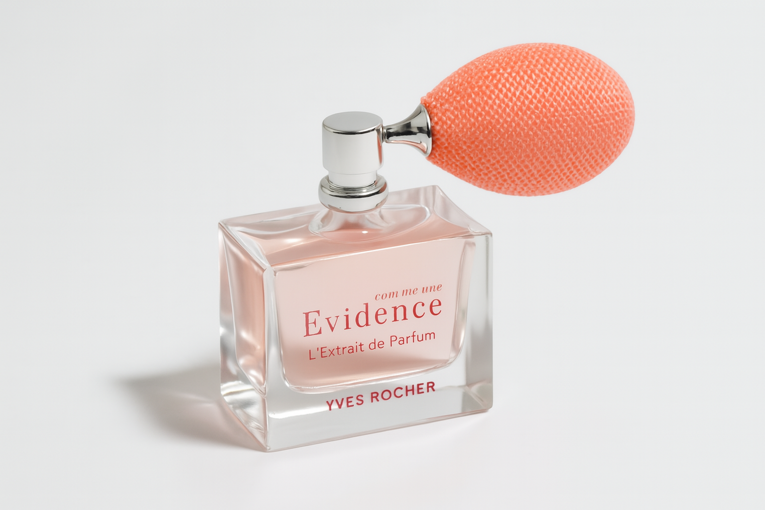 Ein zart roséfarbenes Parfumflakon von Yves Rocher „Comme une Evidence – L’Extrait de Parfum“ steht leicht zur Seite geneigt auf einer neutralen weißen Fläche. Das klare Glas ist rechteckig mit sanft geschwungenen Kanten. Die Vorderseite zeigt die rosafarbene Aufschrift, ergänzt durch einen Retro-Zerstäuber mit orangefarbener Textilpumpe. Ein natürlicher, weicher Schatten sorgt für Tiefe.