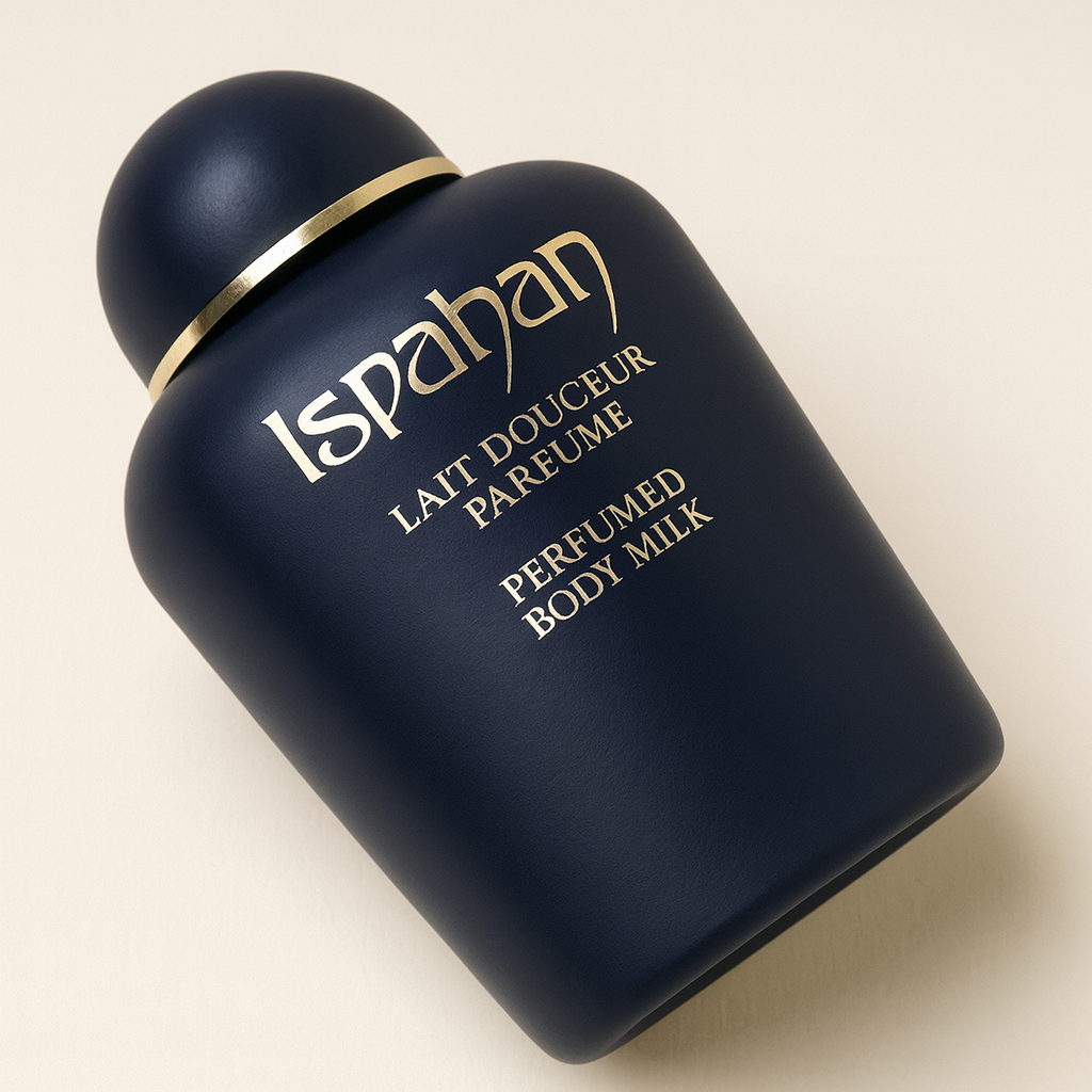 Eine nach hinten geneigte, dunkelblaue Flasche „Ispahan Perfumed Body Milk“ mit matter Oberfläche und goldener Aufschrift. Der runde Verschluss ist ebenfalls dunkelblau und mit einem goldenen Zierband versehen. Die Flasche steht vor einem neutralen, hellen Hintergrund mit weichem Schatten.