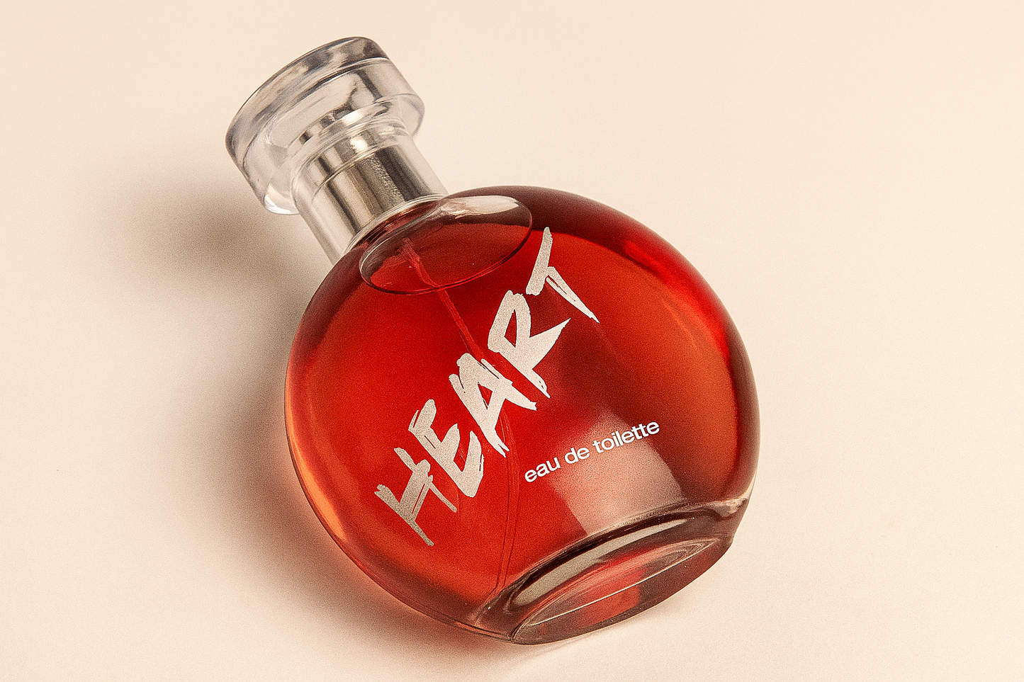 Ein rundes, rotes Parfumflakon mit silbernem Zerstäuber und durchsichtigem Deckel liegt leicht zur Seite geneigt auf einem neutralen beigen Hintergrund. Die Aufschrift „HEART eau de toilette“ ist klar lesbar auf der Vorderseite.