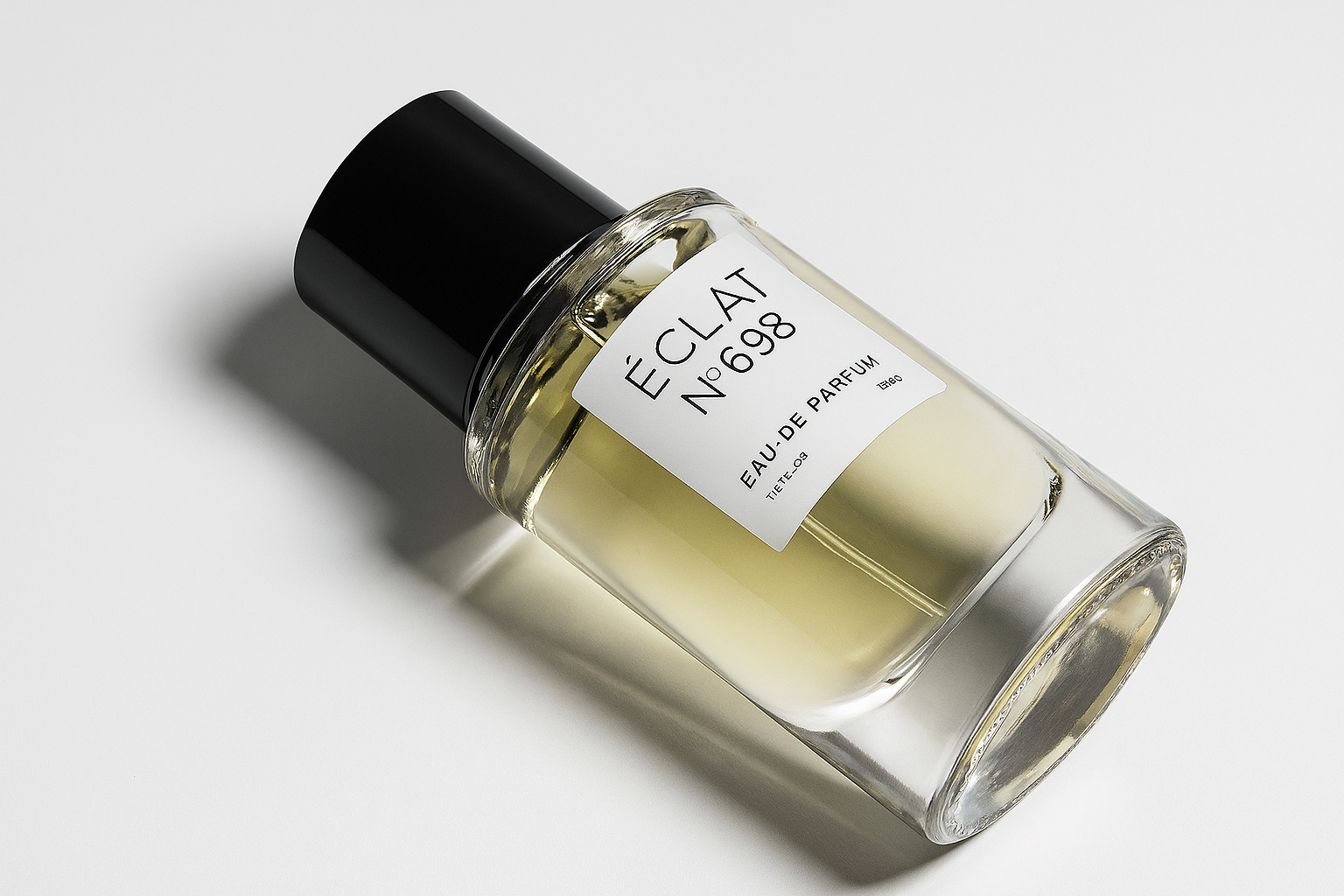 Ein elegantes, transparentes Parfumflakon mit der Aufschrift „ÉCLAT N°698“ liegt leicht seitlich geneigt auf einer neutralen, weißen Oberfläche. Der schwarze Verschluss ist glänzend, und das Fläschchen wirft einen weichen, natürlichen Schatten.