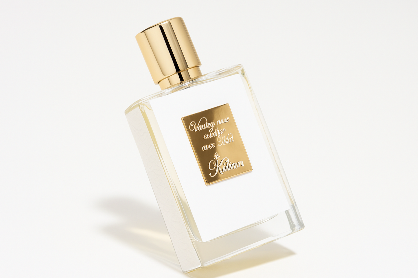 Ein eleganter, leicht nach hinten gekippter Flakon des Parfums "Voulez-vous coucher avec Moi" von Kilian, mit goldfarbener Kappe und goldener Plakette auf der Vorderseite, steht auf einem neutralen, hellen Hintergrund. Der weiche Schatten des Flakons verleiht dem Bild Tiefe.